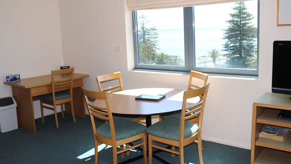 A room with a round wooden table, four wooden chairs with green cushions, a small wooden desk, a trash bin, and a window with a view of trees and the sea.