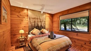 A wooden-paneled bedroom with a bed, a nightstand, a lamp, and a large window offering a view of trees. - Siver Cabin (Kangaroo Valley)