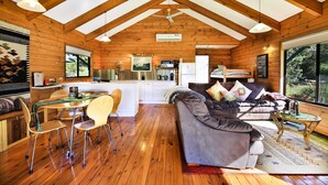 A spacious cabin with a high ceiling, wooden walls, a dining area with chairs and a table, a sofa with cushions, and a large window offering a view of the outdoors. - Siver Cabin (Kangaroo Valley)