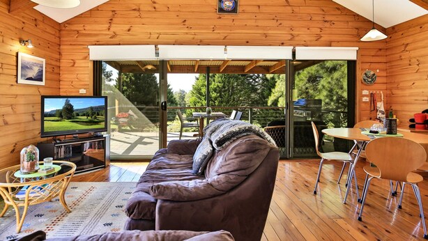 A cozy cabin interior with a wooden ceiling, a large TV, a sofa, and a dining area with chairs and a table. - Siver Cabin (Kangaroo Valley)