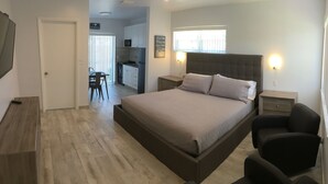 Een slaapkamer met een groot bed, nachtkastjes, een bureau en een stoel. Achterin is een kitchenette te zien.