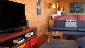 Ein Wohnzimmer mit einem großen Flachbildfernseher, einem Couchtisch mit Schachfiguren, einer Couch mit gestreiften Kissen und einem Bücherregal voller Bücher.