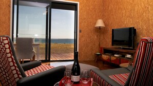 Ein Zimmer mit einer großen Glasschiebetür, die Blick auf einen Strand bietet, einem Fernseher und zwei Stühlen. Auf dem Tisch stehen eine Weinflasche und Gläser.