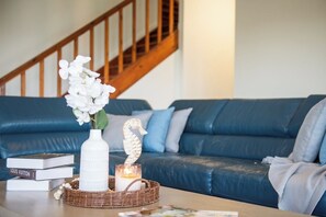 Un salon avec un canapé en cuir bleu, un vase blanc rempli de fleurs, une bougie et un panier en osier posé sur une table basse en bois.