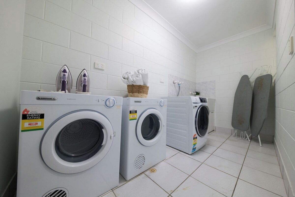 A laundry area with three washing machines, a basket of folded towels, and a pair of ironing boards.