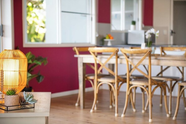 A dining area with wicker chairs and a table, a wicker lamp on a side table, and a window with a view of greenery.