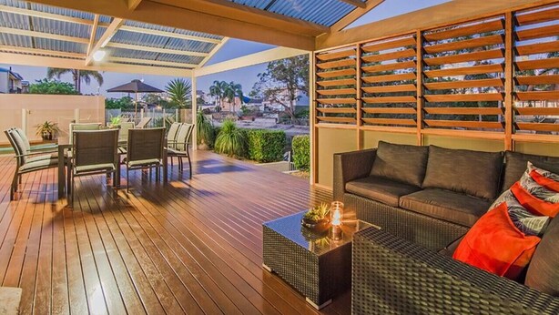 Um pátio externo com deck de madeira, área de estar com sofá e cadeiras, e vista da paisagem ao redor.