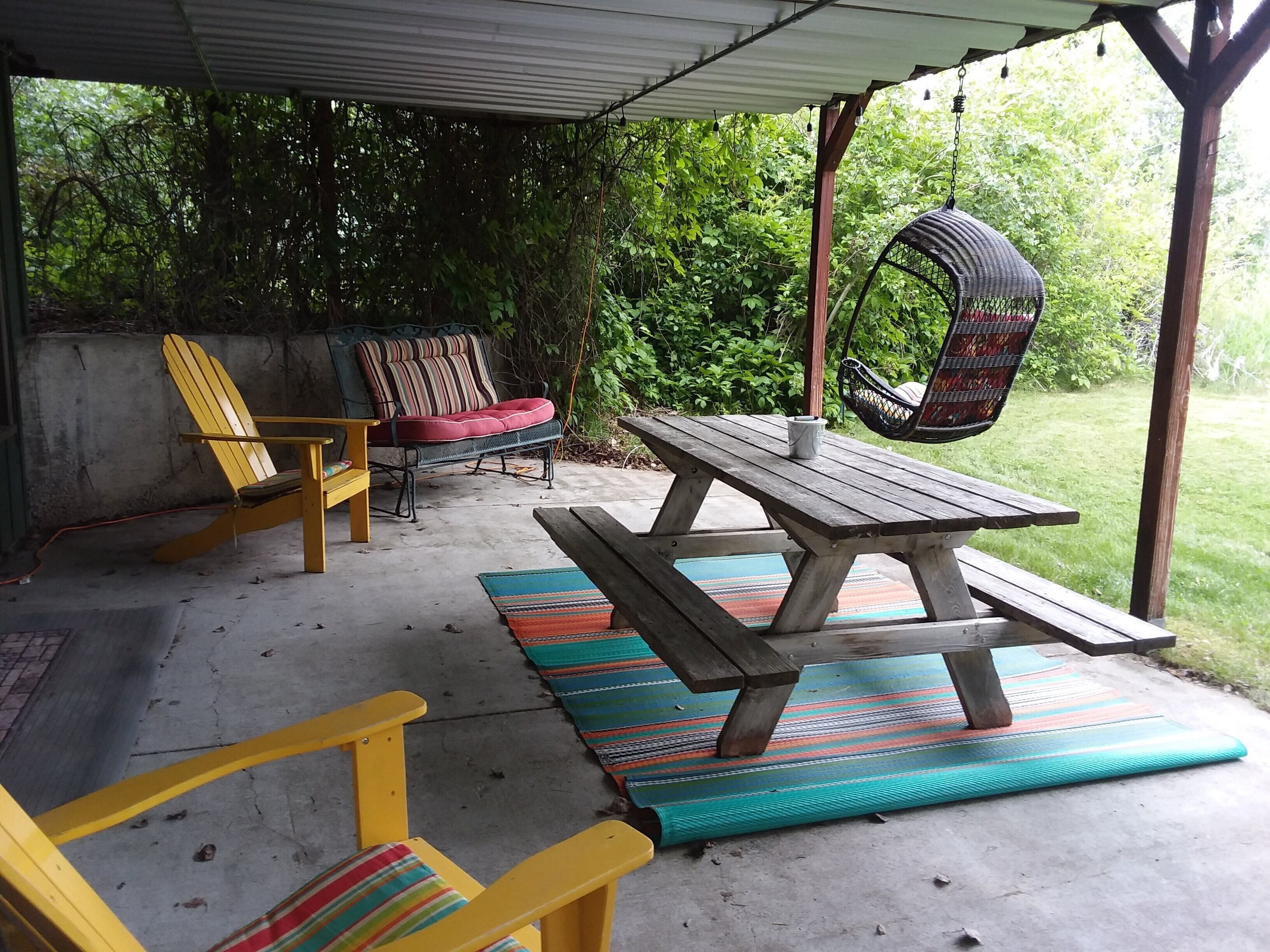 An outdoor patio with a wooden picnic table, yellow chairs, a cushioned chair, and a hanging chair.