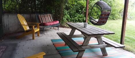 An outdoor patio with a wooden picnic table, yellow chairs, a cushioned chair, and a hanging chair.