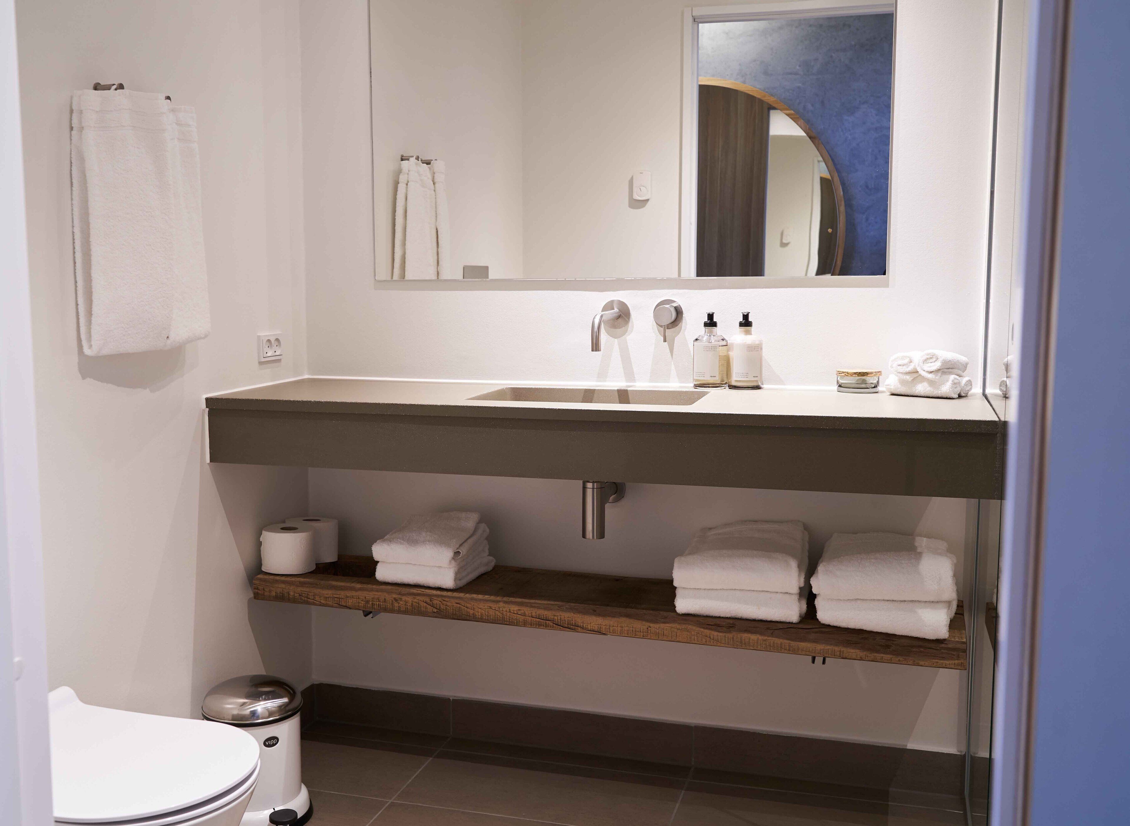 A bathroom with a white sink, a mirror, a toilet, and shelves with folded towels.