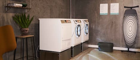 Een wasruimte met drie voorladers wasmachines en drogers, een plant in pot, een stoel en een prikbord met instructies.