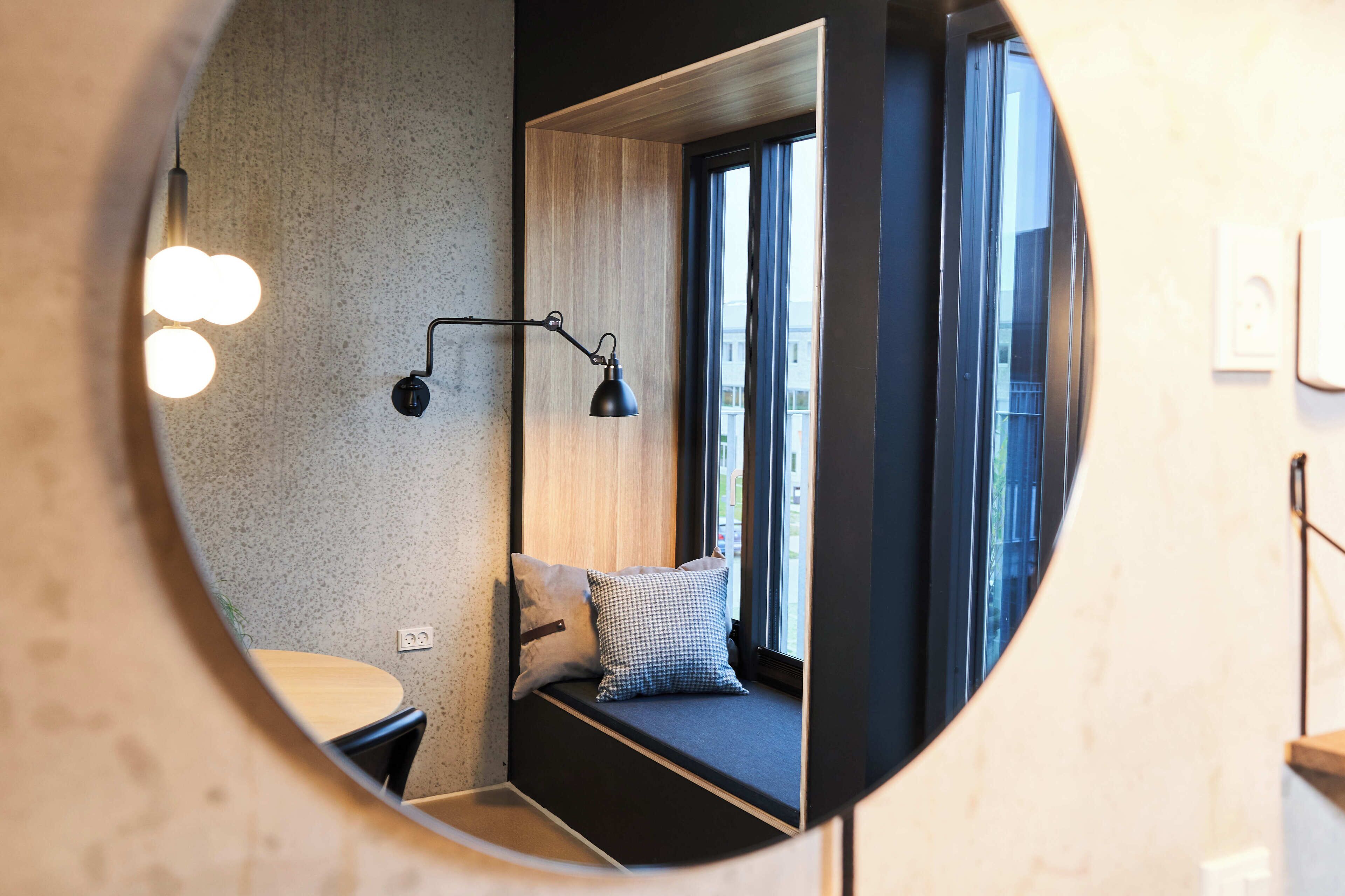 Un espejo circular que refleja una habitación con una cama, una mesita de noche, una lámpara y una ventana con cortinas.