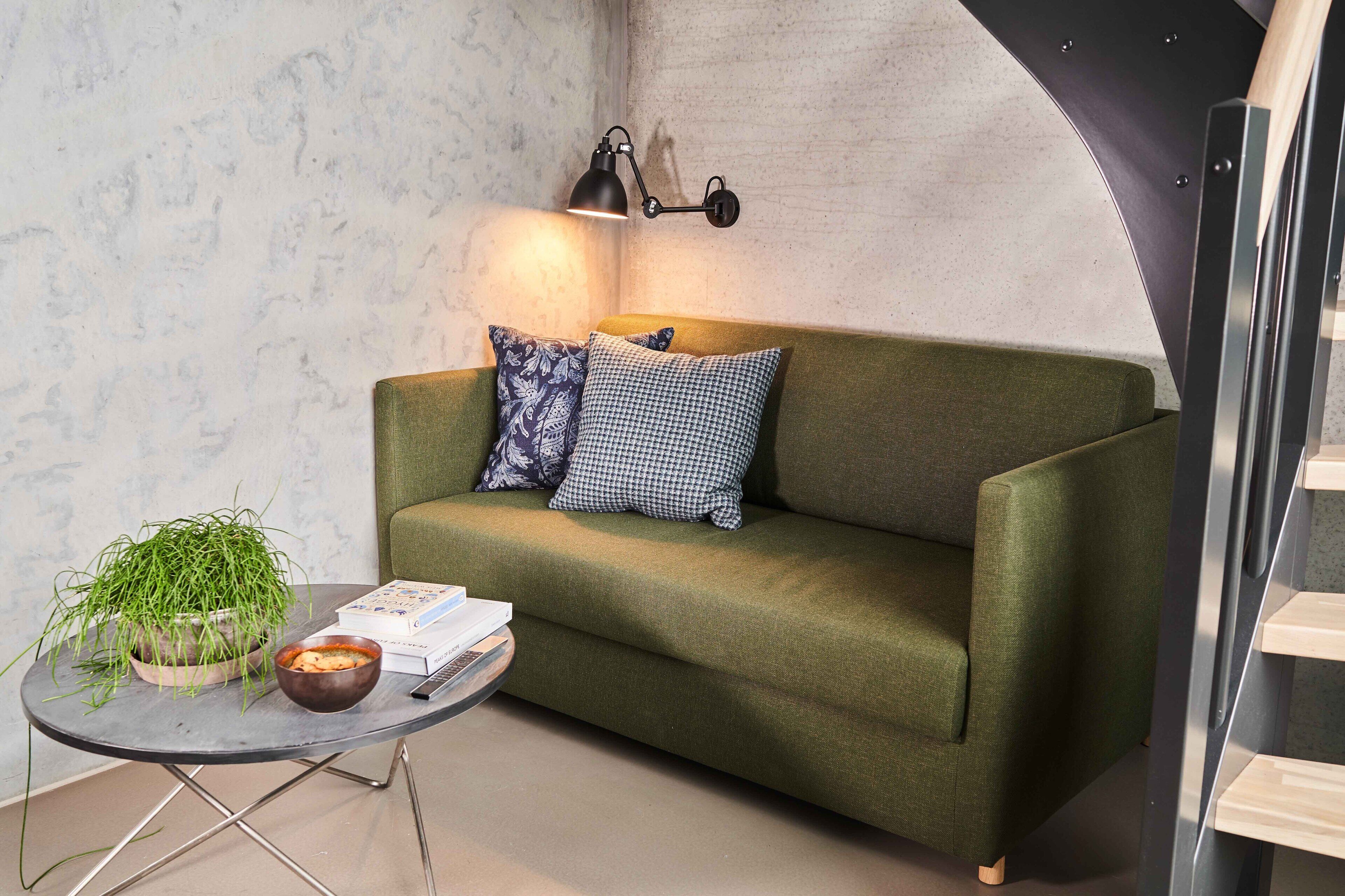 Un sofá verde con cojines estampados, una pequeña mesa redonda con una planta y libros, y una lámpara fijada a la pared.