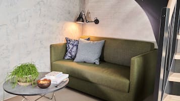 A green sofa with patterned cushions, a small round table with a plant and books, and a wall-mounted lamp.