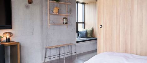 A modern bedroom with a bed, a wooden desk with a lamp, a TV, and a window with a view.