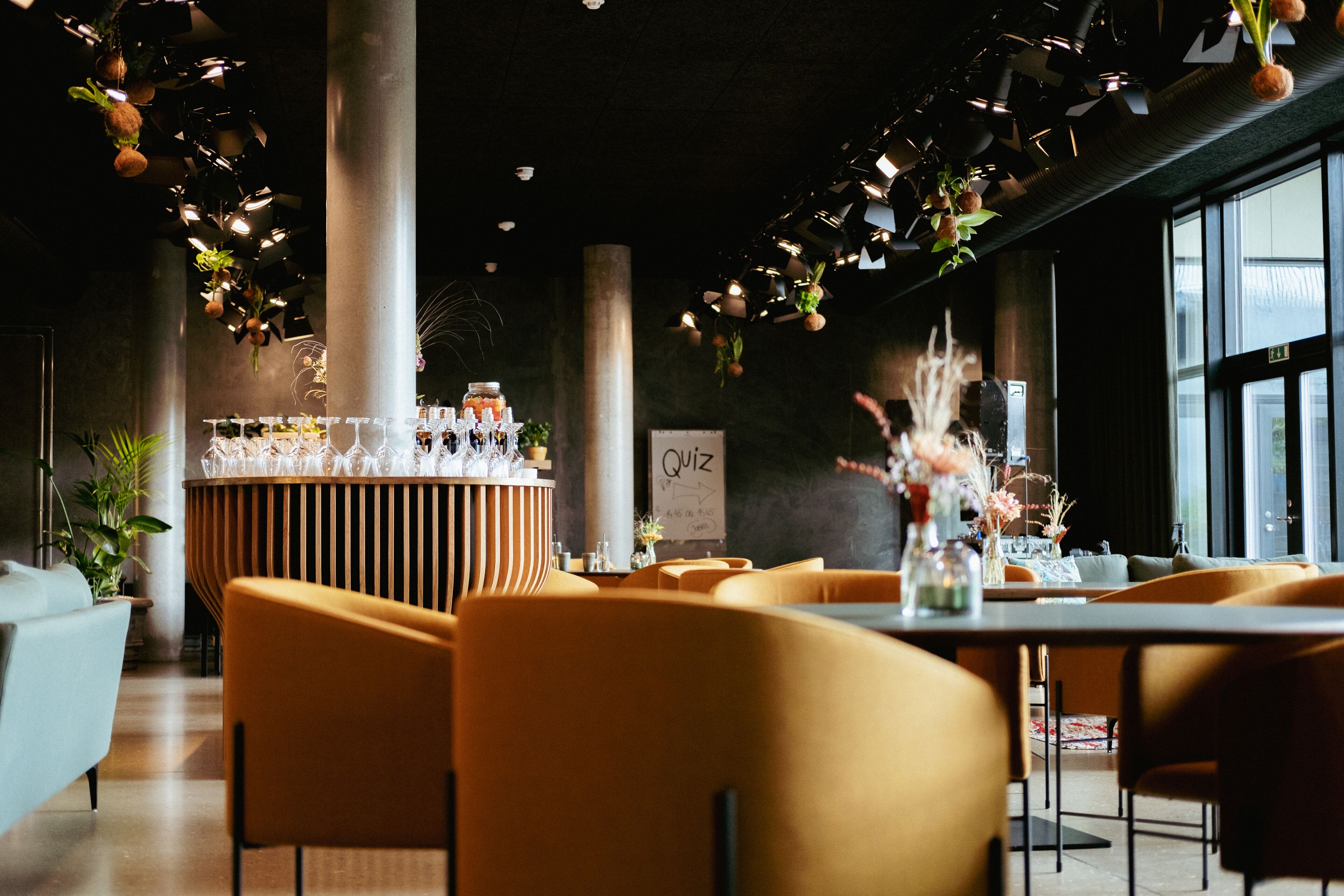 Et moderne restaurantmiljø med en central bar, orange siddepladser og dekorative hængende planter.