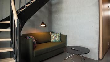 A modern interior with a curved staircase, a green sofa with a colorful pillow, a small round table, and a wall-mounted light fixture.