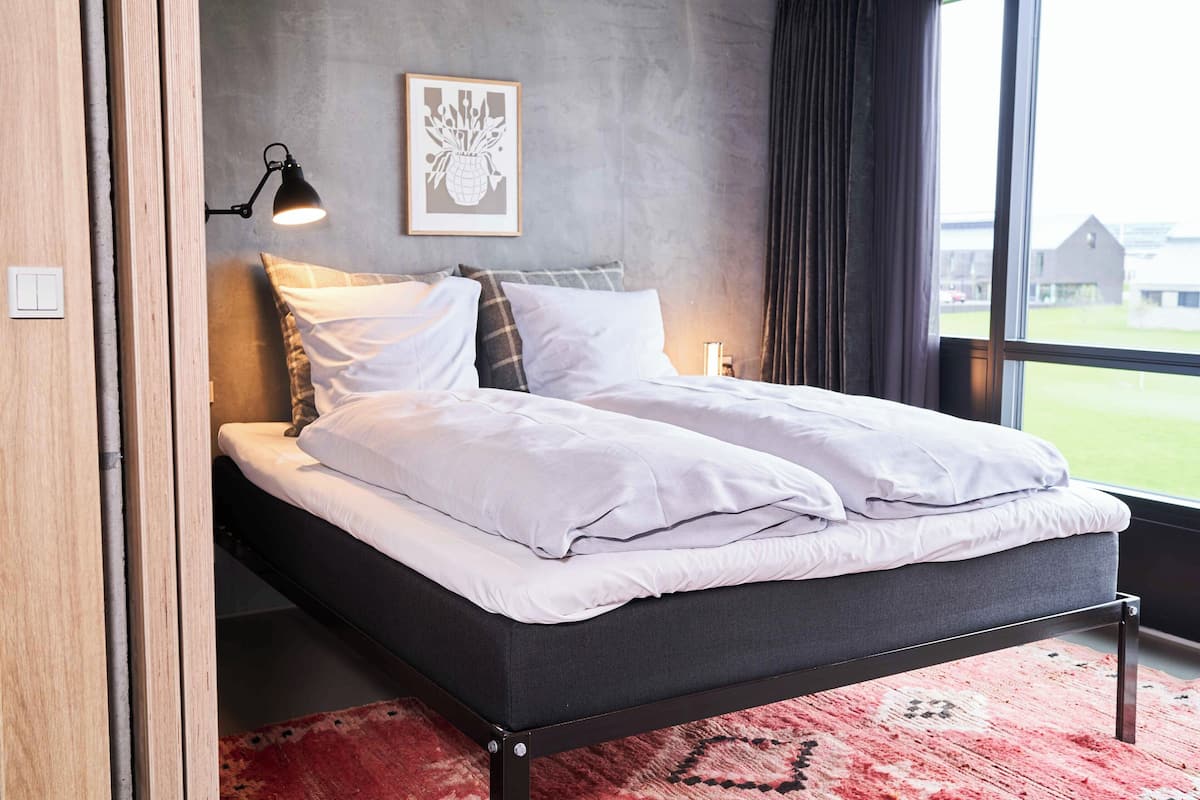 A modern bedroom with a bed, a wall-mounted lamp, and a window with a view of a building.