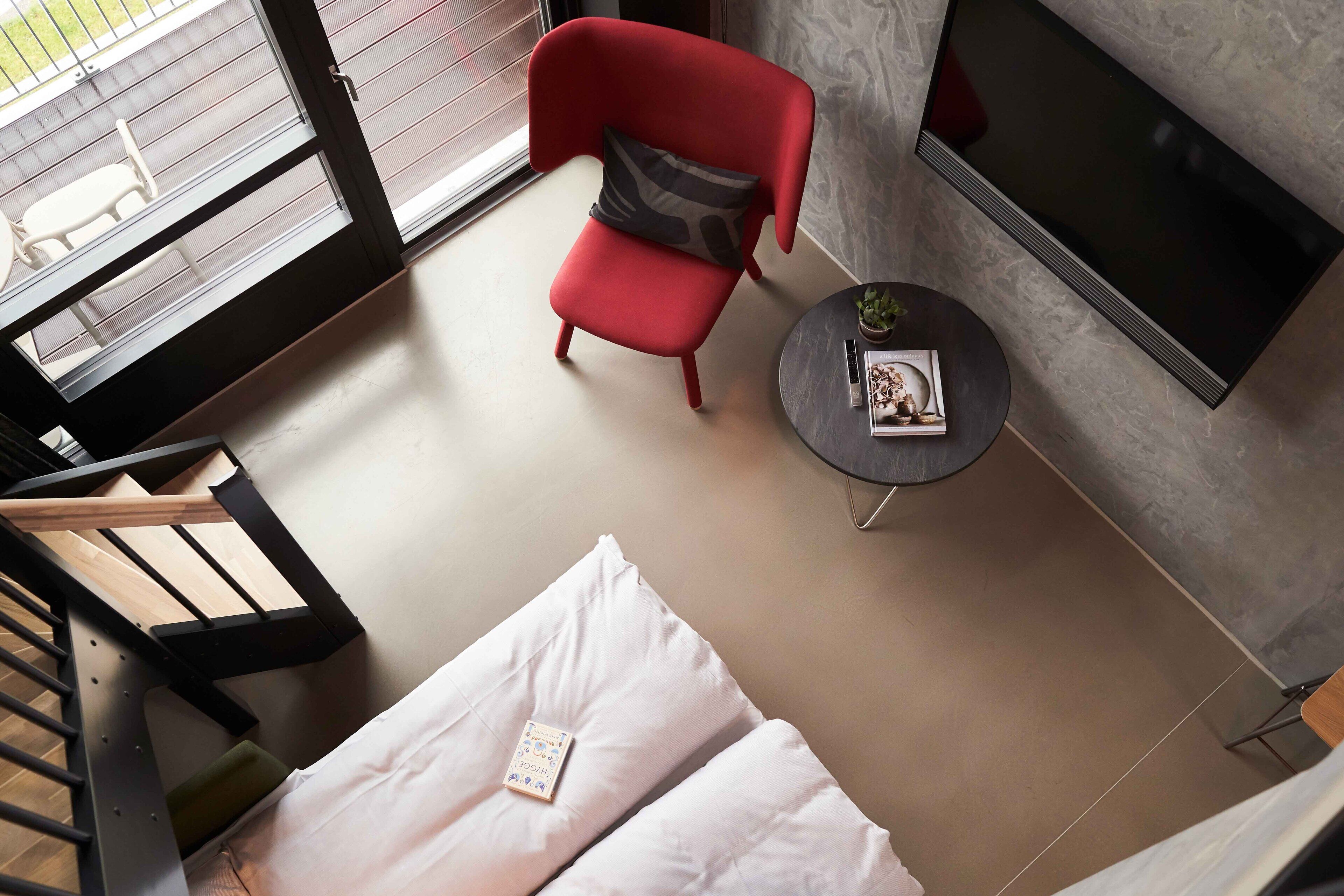 A modern room with a red chair, a small round table with a book and a plant, a bed with white bedding, and a TV mounted on the wall.