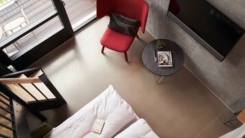 Une chambre moderne avec un fauteuil rouge, une petite table ronde sur laquelle se trouvent un livre et une plante, un lit avec une literie blanche, et un téléviseur fixé au mur.