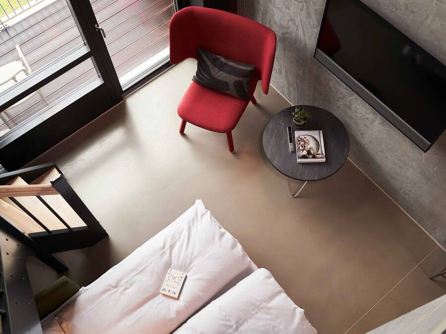 Une chambre moderne avec un fauteuil rouge, une petite table ronde sur laquelle se trouvent un livre et une plante, un lit avec une literie blanche, et un téléviseur fixé au mur.