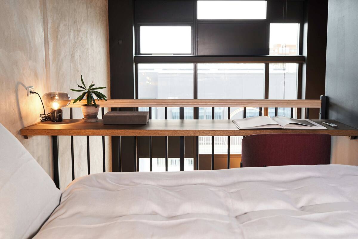 A bedroom with a bed, a bedside table with a lamp, a plant, and an open book.
