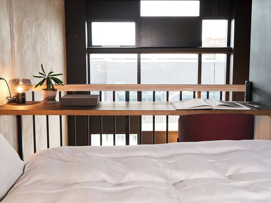 A bedroom with a bed, a bedside table with a lamp, a plant, and an open book.