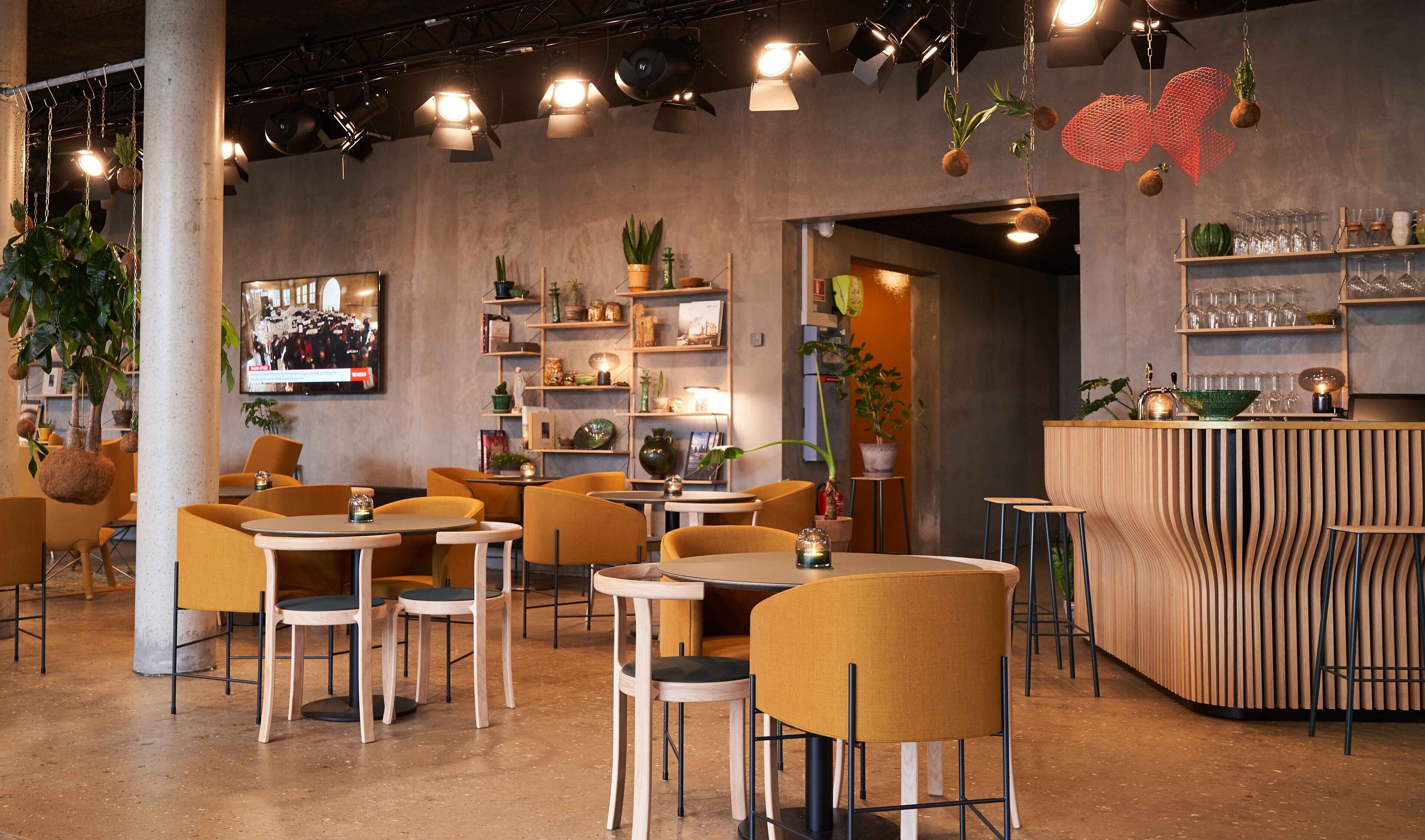 Un caffè moderno con tavoli rotondi e sedie gialle, scaffali con piante e oggetti decorativi, e una zona bar con sgabelli.