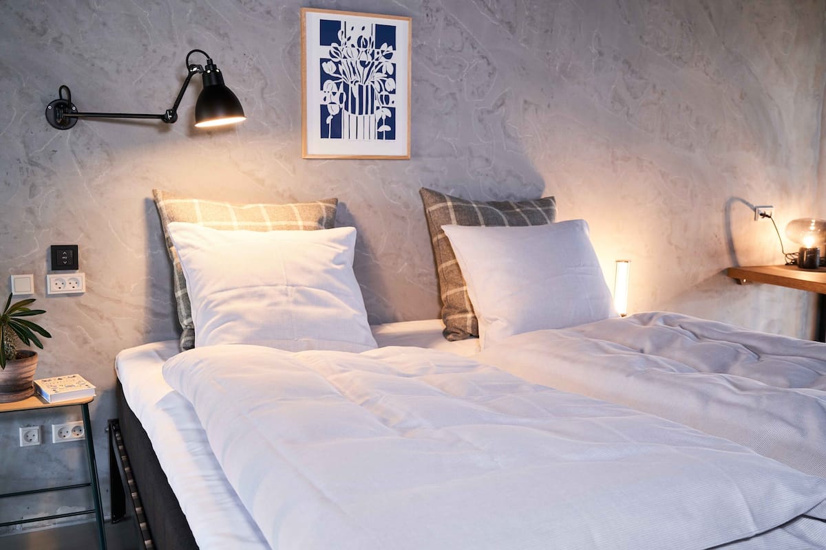 A neatly made bed with white linens, a bedside table with a lamp, and a wall-mounted artwork.