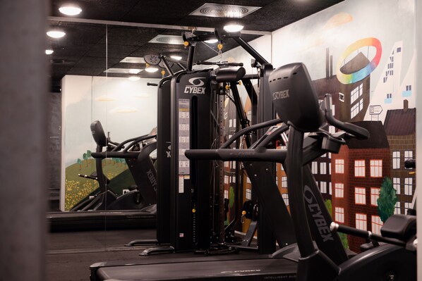 Une salle de sport équipée de machines Cybex, ornée d’une fresque représentant un paysage urbain et dotée d’un mur miroir.