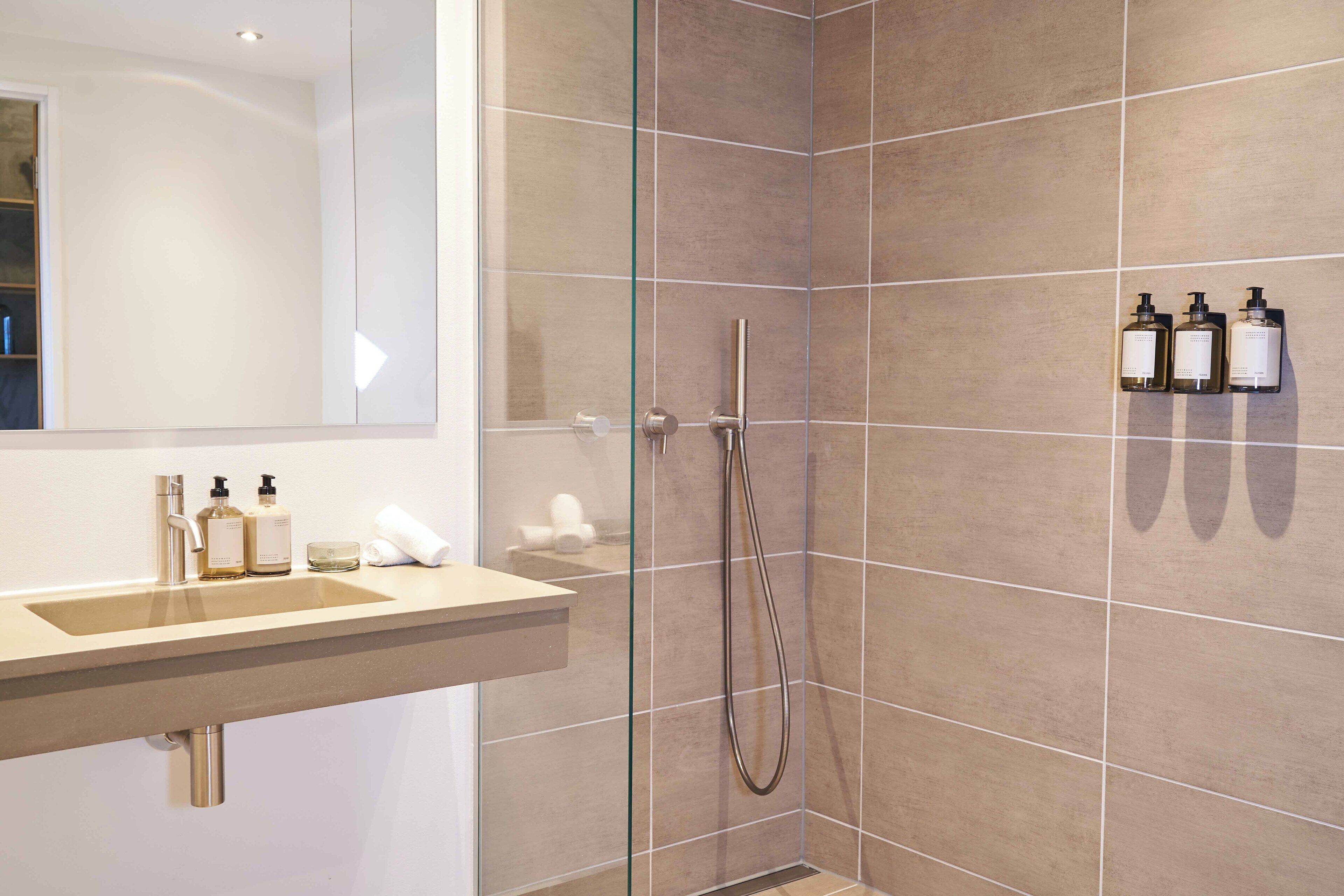 Un baño moderno con ducha revestida de baldosas beiges, un lavabo con dos botellas de jabón líquido y un espejo.
