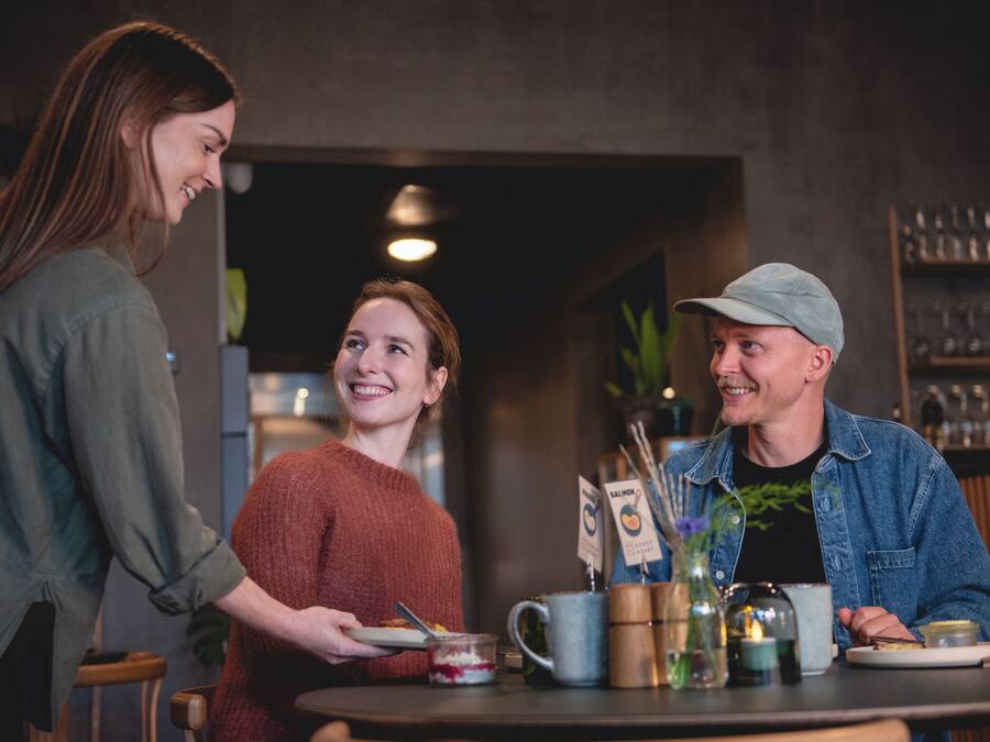 Un grupo de personas sentadas alrededor de una mesa, con un menú, una cafetera y un jarrón con una planta.
