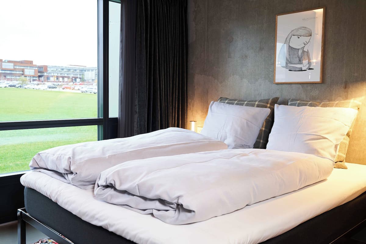 A hotel room with a large window, a bed with white linens, a framed artwork on the wall, and a view of a building and parking lot outside.