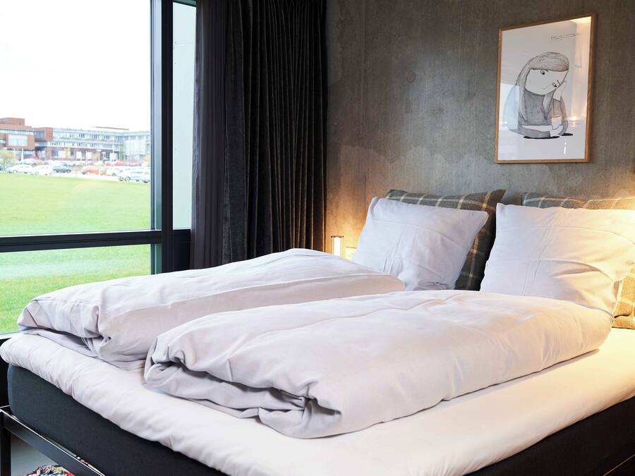 Ein Hotelzimmer mit großem Fenster, einem Bett mit weißer Bettwäsche, einem gerahmten Kunstwerk an der Wand und Blick auf ein Gebäude und einen Parkplatz.