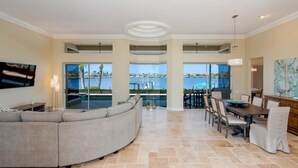 Ein großzügiger Wohnbereich mit einer gebogenen, grauen Couch, einem Esstisch mit Stühlen und Blick auf Pool und Meer durch große Fenster.