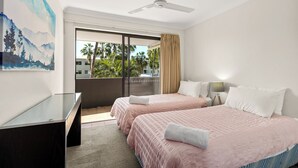 Ein Hotelzimmer mit zwei Betten, einem Schreibtisch, einem Deckenventilator und einem großen Fenster mit Blick auf Palmen.