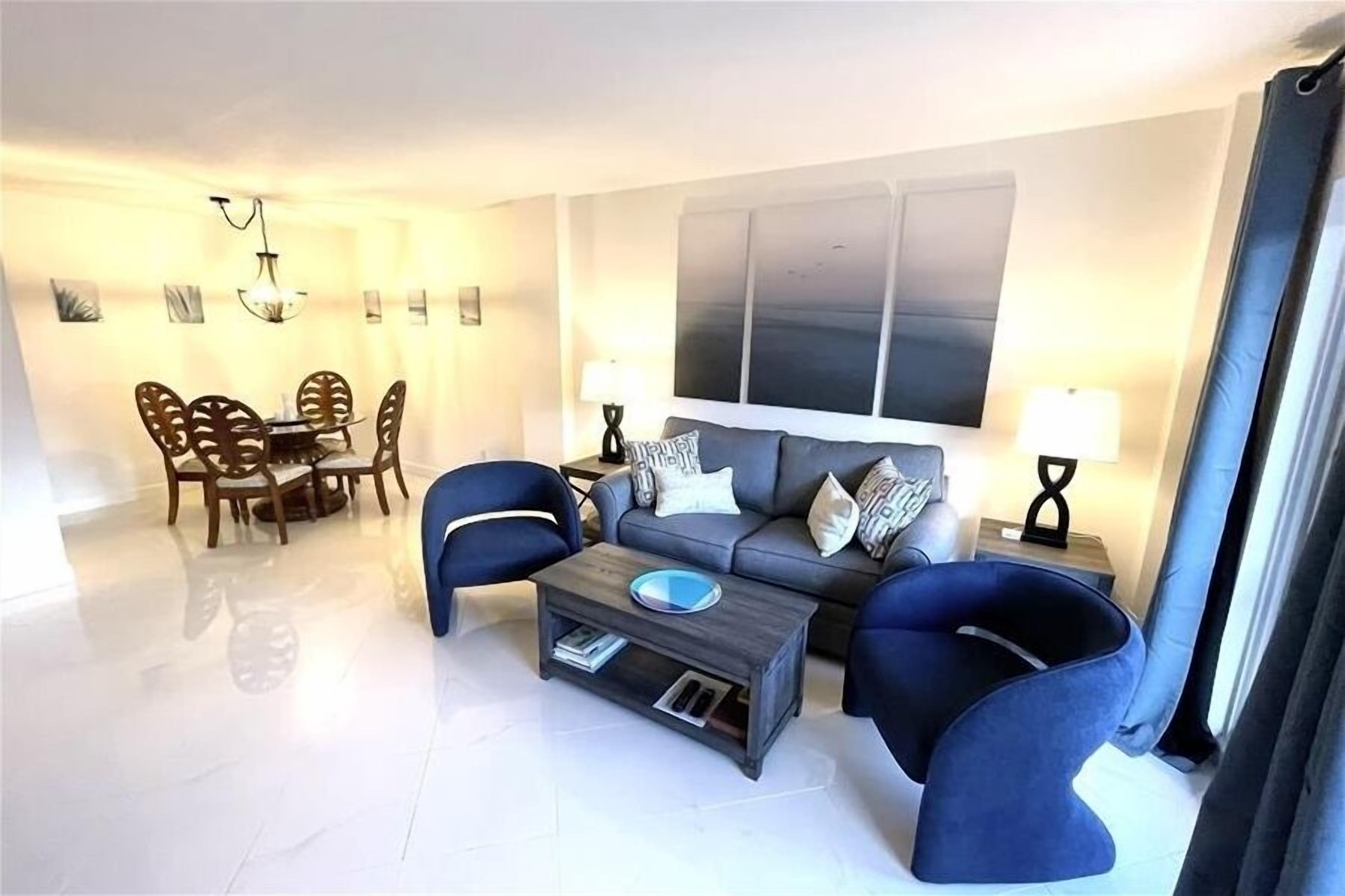 A modern living room with a grey sofa, two blue armchairs, a coffee table, and a dining area with a wooden table and chairs.