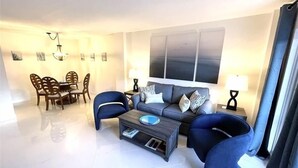 Una sala de estar moderna con un sofá gris, dos sillones azules, una mesa de centro y un comedor con una mesa y sillas de madera.