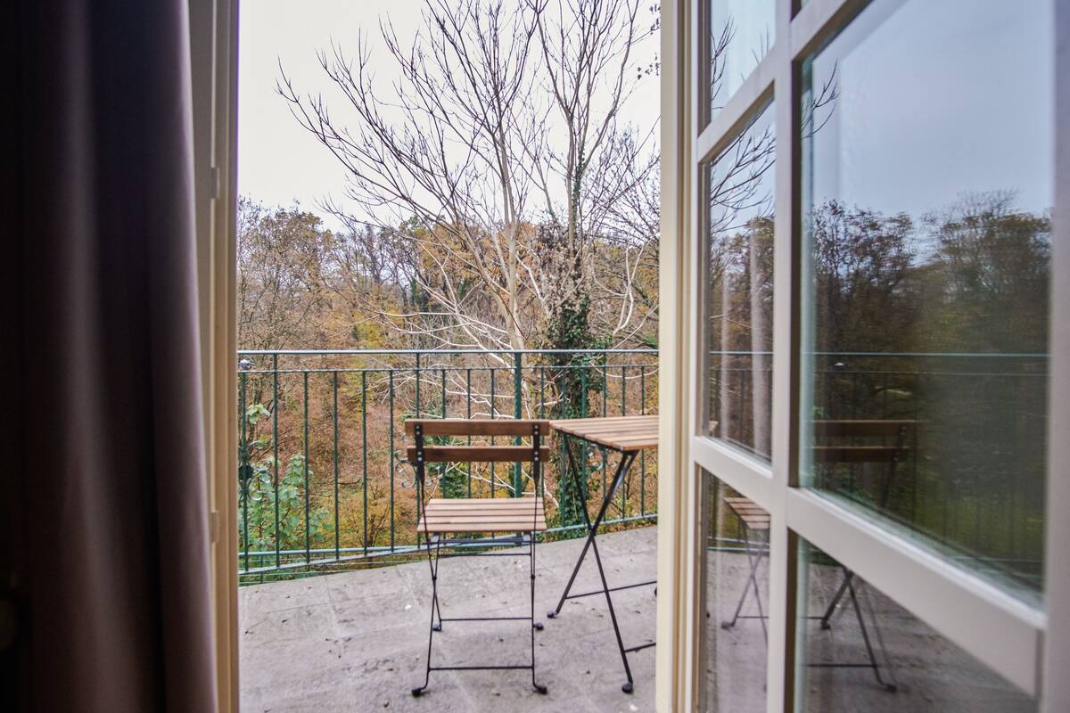 Ein Balkon mit Holztisch und -stühlen, einem Metallgeländer und Blick auf Bäume und ein Gebäude.