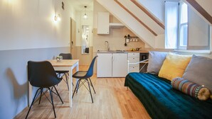 A compact room with a dining table, chairs, a bed with a colorful blanket, a kitchenette, and a window with curtains. - Peacock Boutique Apartments (Zagreb)