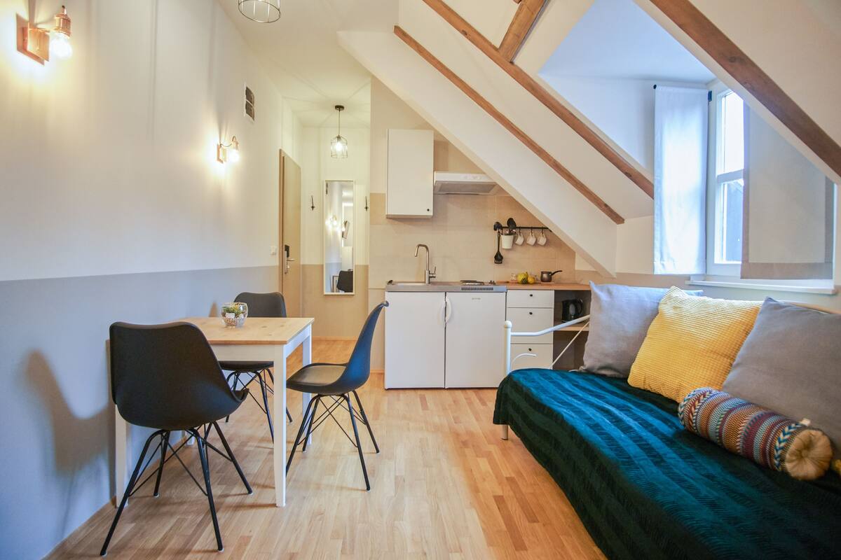 Ein kleines Zimmer mit Esstisch, Stühlen, einem Bett mit bunter Decke, einer Küchenzeile und einem Fenster mit Vorhängen.