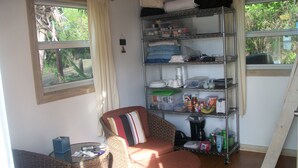 A room with wicker furniture, a metal shelf unit with various items, and a window with curtains. - Tiny House Vacation, Bayfront with private beach area!  On two secluded acres! (Dunmore Town)
