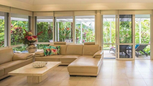 A spacious living area with a beige sectional sofa, a coffee table, and a view of lush greenery through large windows. - Port Douglas Mirage Villa 123 (Port Douglas)
