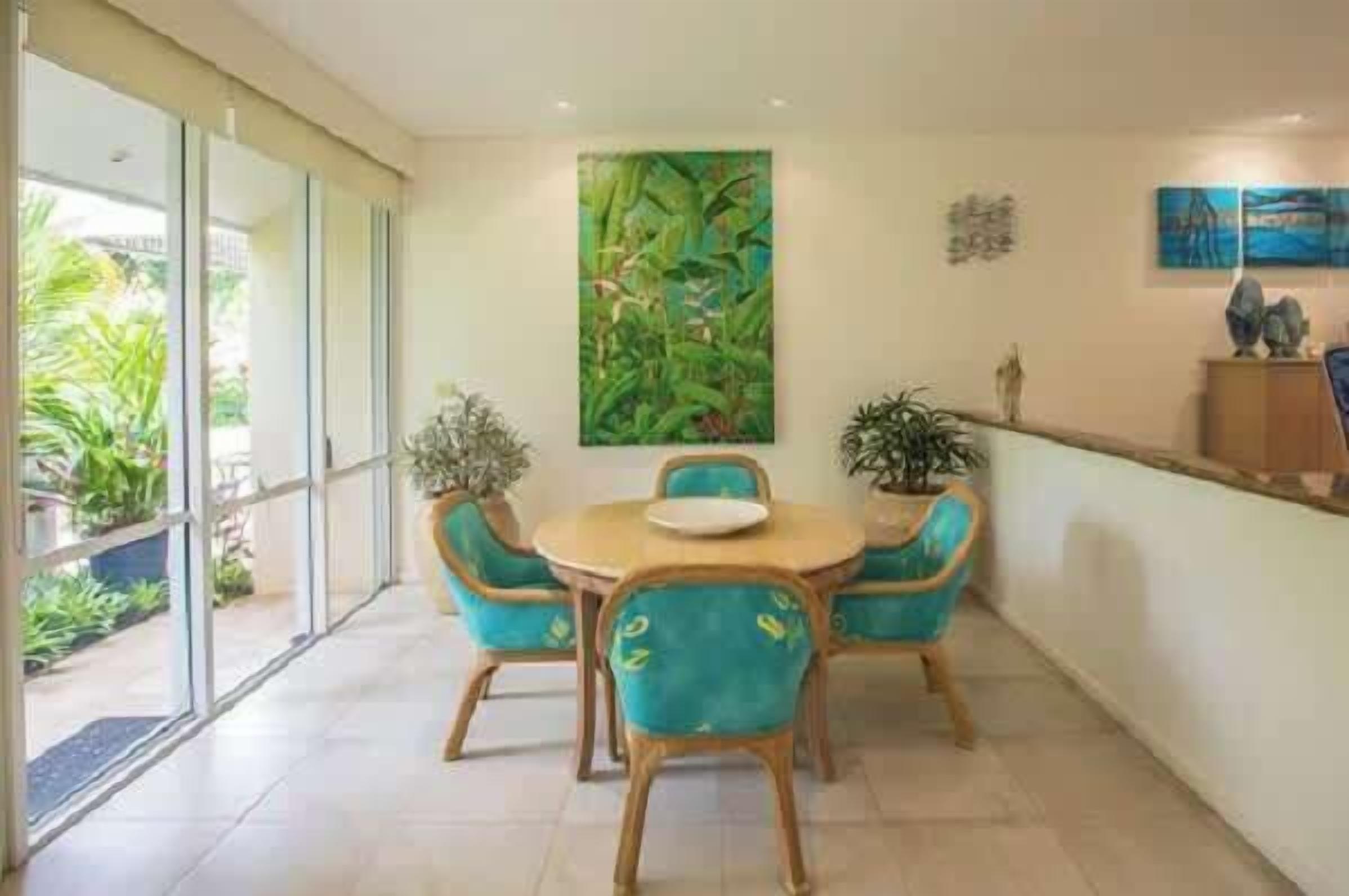 Un comedor con una mesa redonda y cuatro sillas, un cuadro grande en la pared y vistas al exterior con vegetación.