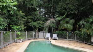 Ein Swimmingpool mit zwei weißen Liegestühlen, umgeben von einem Zaun und üppiger Bepflanzung.