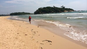 A sandy beach with a person walking towards the water, a small island with greenery in the background, and waves gently crashing onto the shore. - Tara Garden Tropical Villa with Private Pool & Friendly Staff Near Beach (Dharga Town)