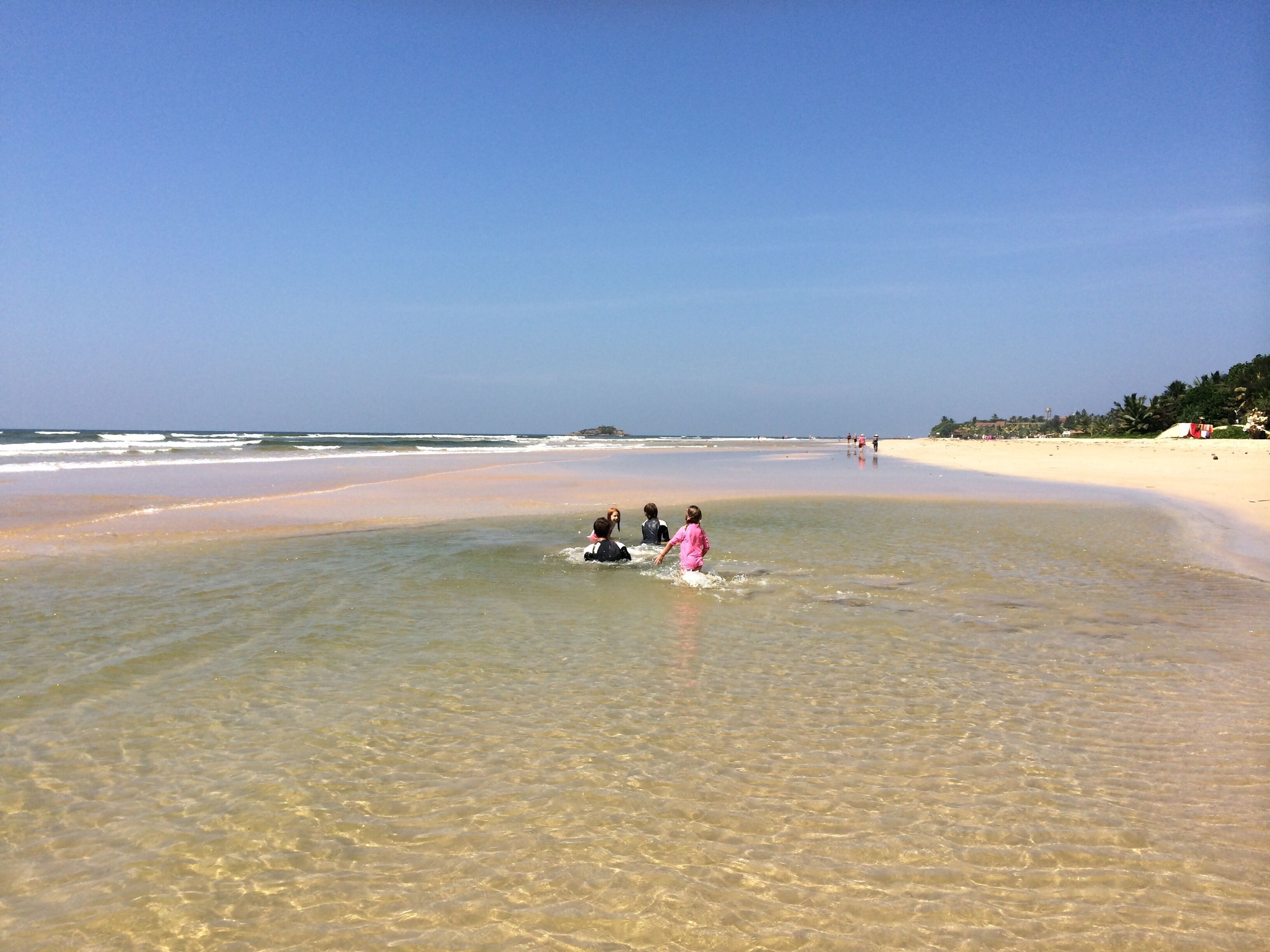 A beach with clear shallow waters, a sandy shore, and a few people in the distance.