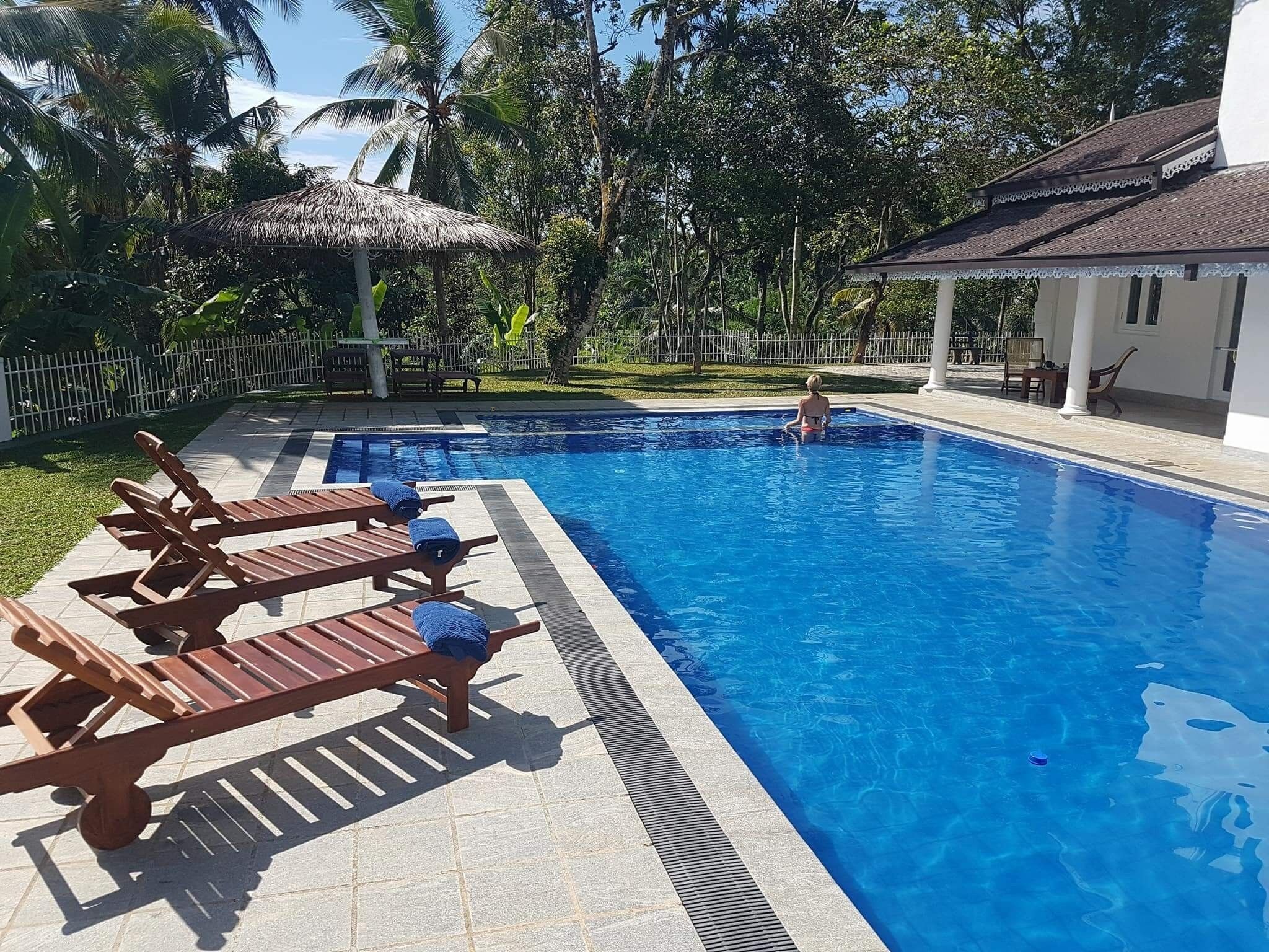 A swimming pool with lounge chairs, a thatched roof structure, and a white building with a tiled roof.
