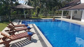 A swimming pool with lounge chairs, a thatched roof structure, and a white building with a tiled roof.