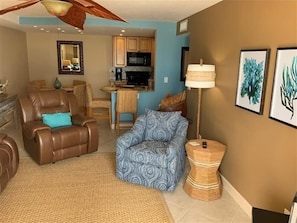 A living room with a brown leather sofa, a patterned blue armchair, and a small round table with a lamp. There is a kitchen in the background with wooden cabinets and a microwave.